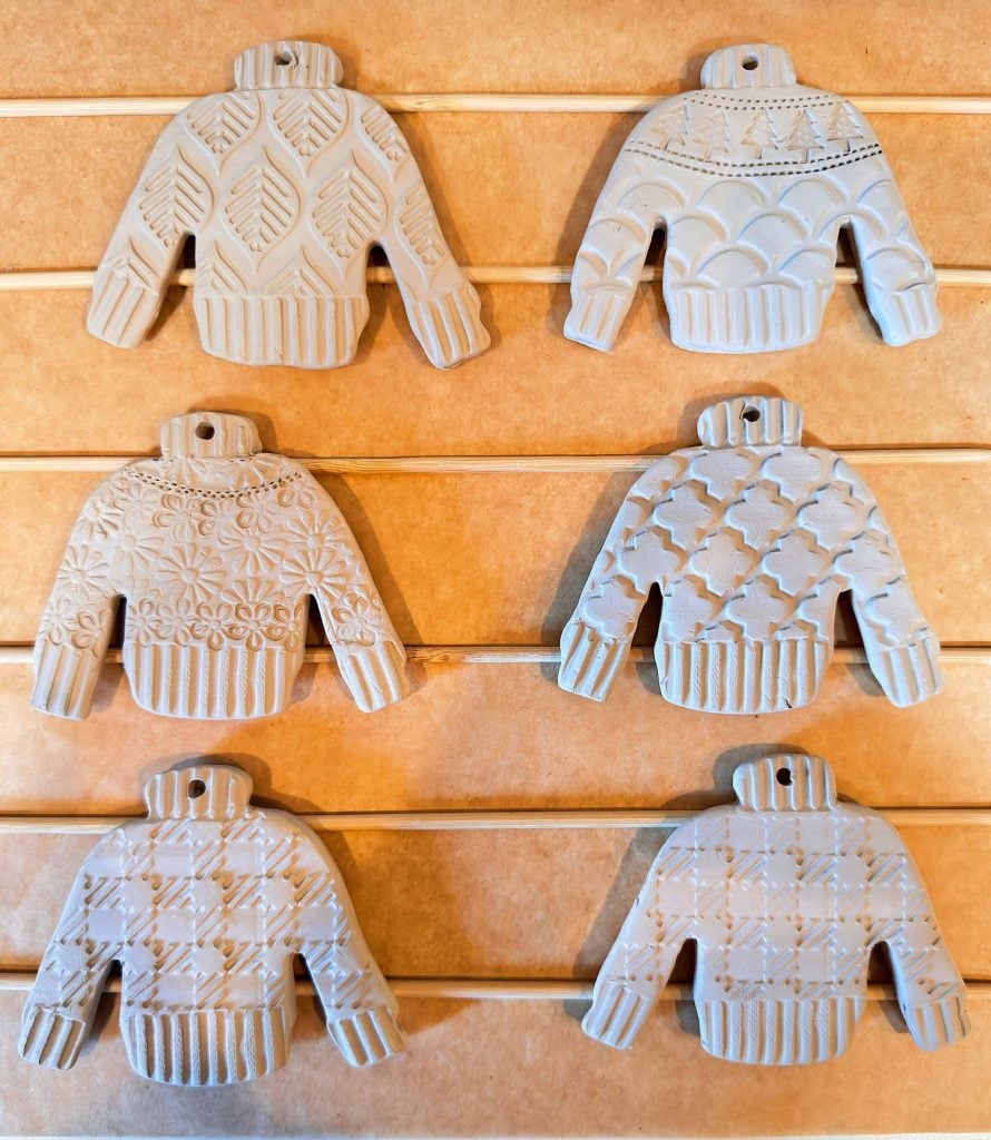 Six ceramic ornaments shaped like sweaters, each with a different textured pattern, are displayed in two rows on a wooden background.