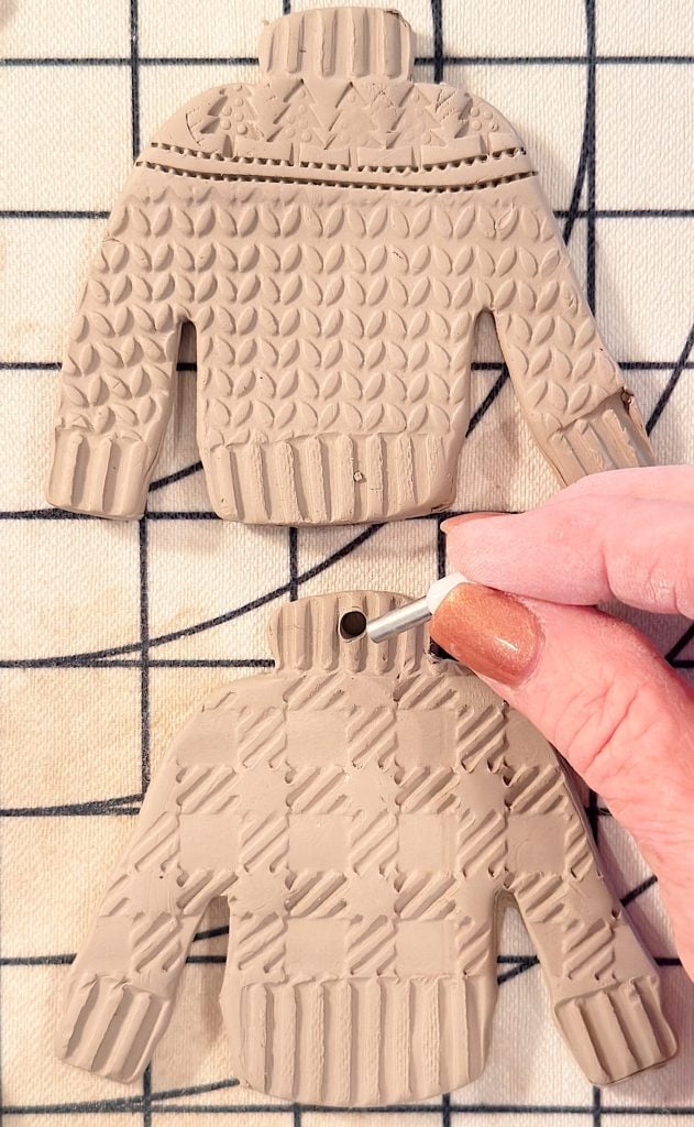 Two textured clay sweater ornaments on a grid surface; a hand uses a tool to punch a hole in the collar of the lower ornament.