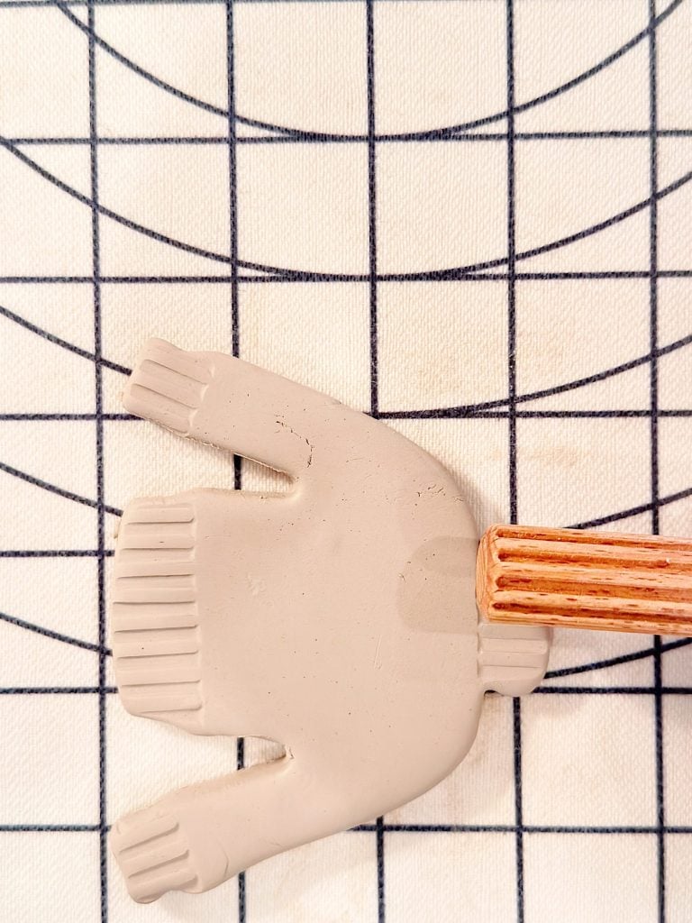 A flat piece of light gray clay shaped like a sweater is pressed by a wooden tool on a gridded fabric surface.