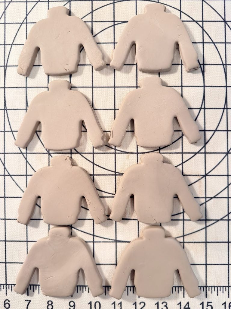 Eight clay pieces shaped like sweaters are arranged in two columns on a gridded cutting mat with measurement markings.