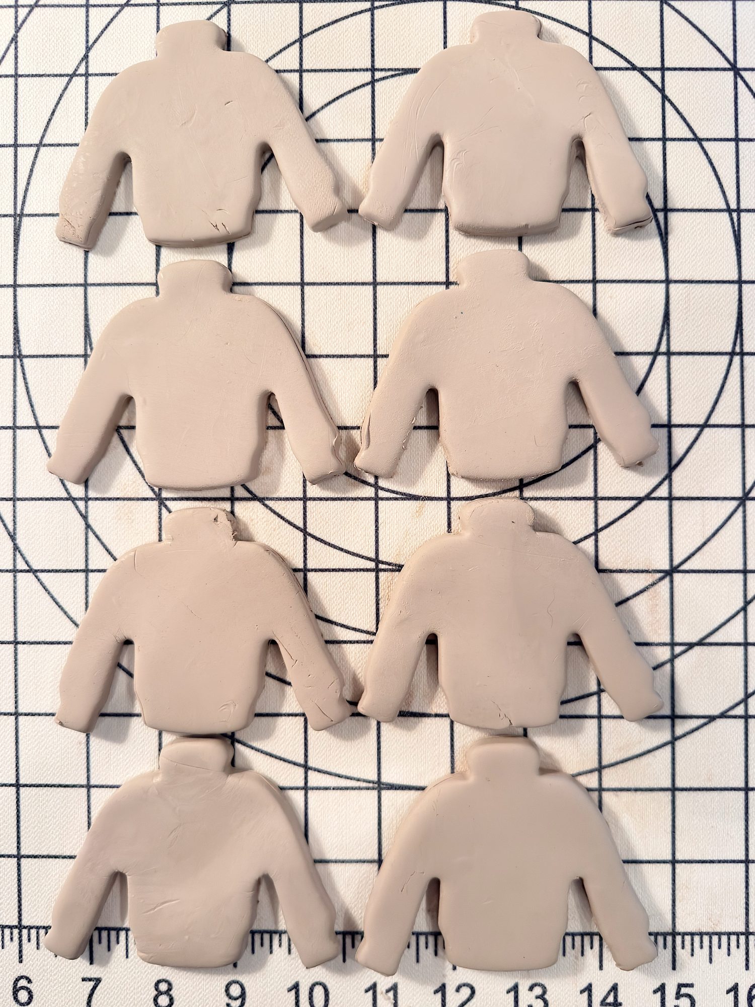 Eight clay pieces shaped like sweaters are arranged in two columns on a gridded cutting mat with measurement markings.