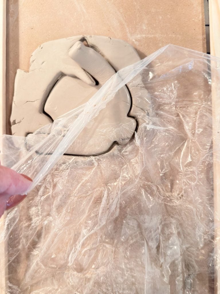 A hand lifts plastic wrap from a slab of clay on a wooden surface, revealing an outlined shape cut into the clay.