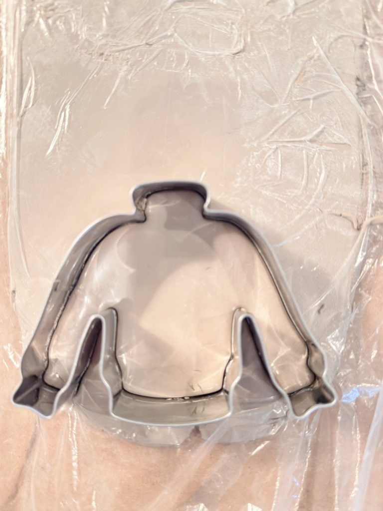 A metal cookie cutter in the shape of a jacket is placed on a sheet of clear plastic wrap.