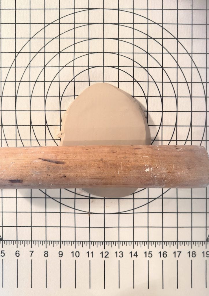 A wooden rolling pin flattening a piece of beige clay on a gridded and circular measurement mat.