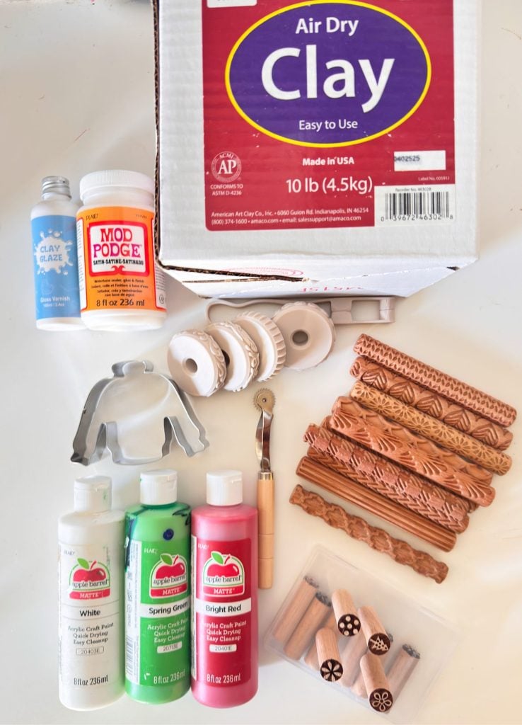 A box of air dry clay with bottles of paint, Mod Podge, clay glaze, patterned rolling pins, cutters, a clay tool, and stamps arranged on a white surface.