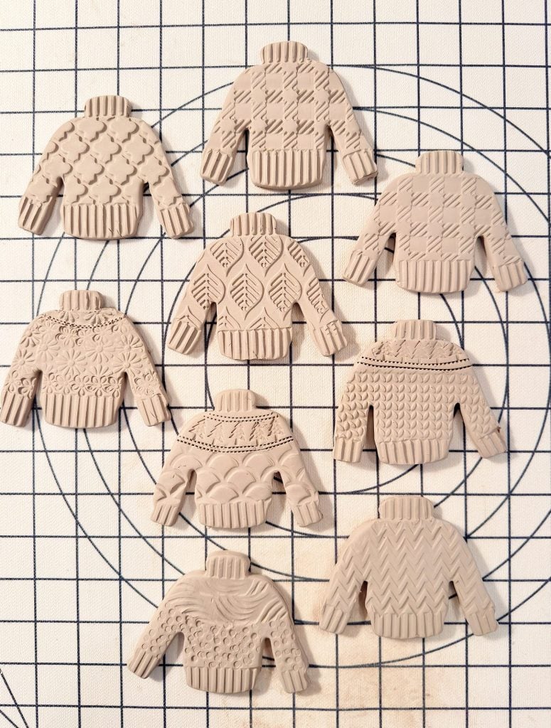 Eight clay sweater figurines with various textured patterns arranged on a grid-lined surface.