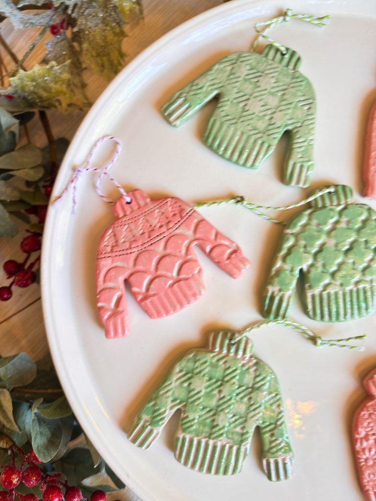 Decorative sweater-shaped ornaments in green and pink with textured patterns are arranged on a white plate, each with a string for hanging.