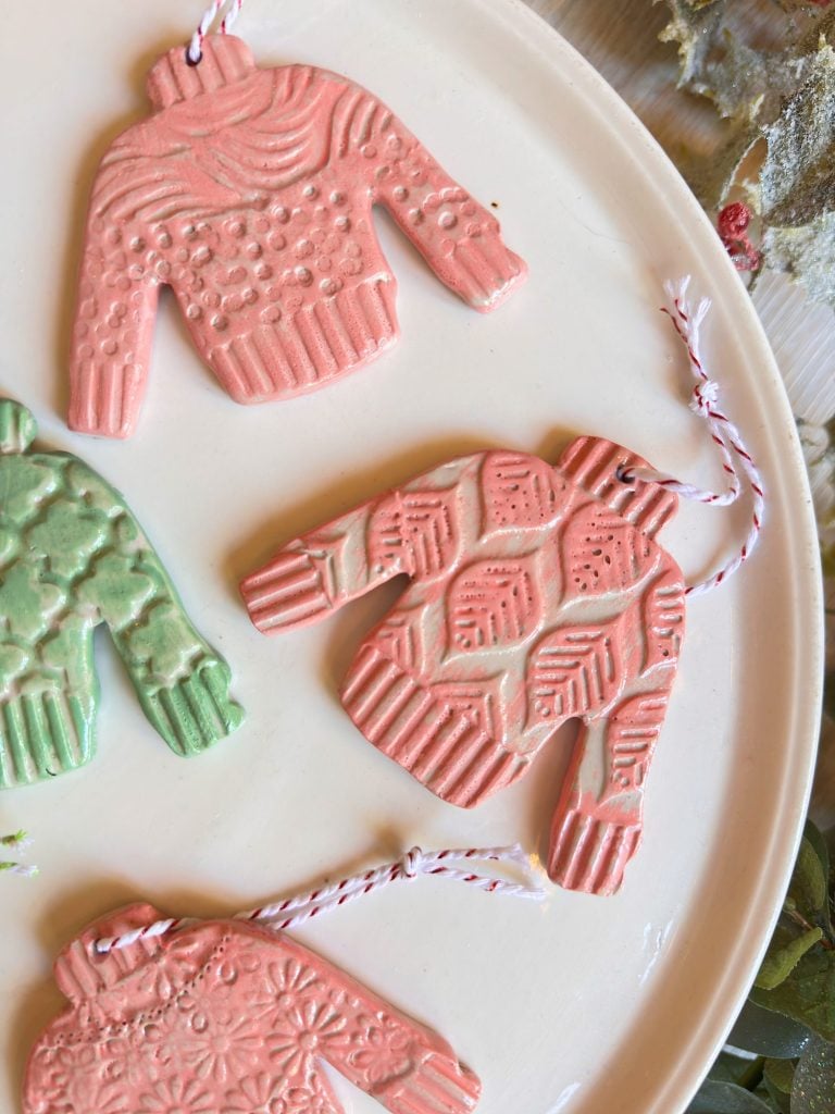 Four sweater-shaped ornaments with textured patterns and pastel colors are displayed on a white plate, each with a string for hanging.