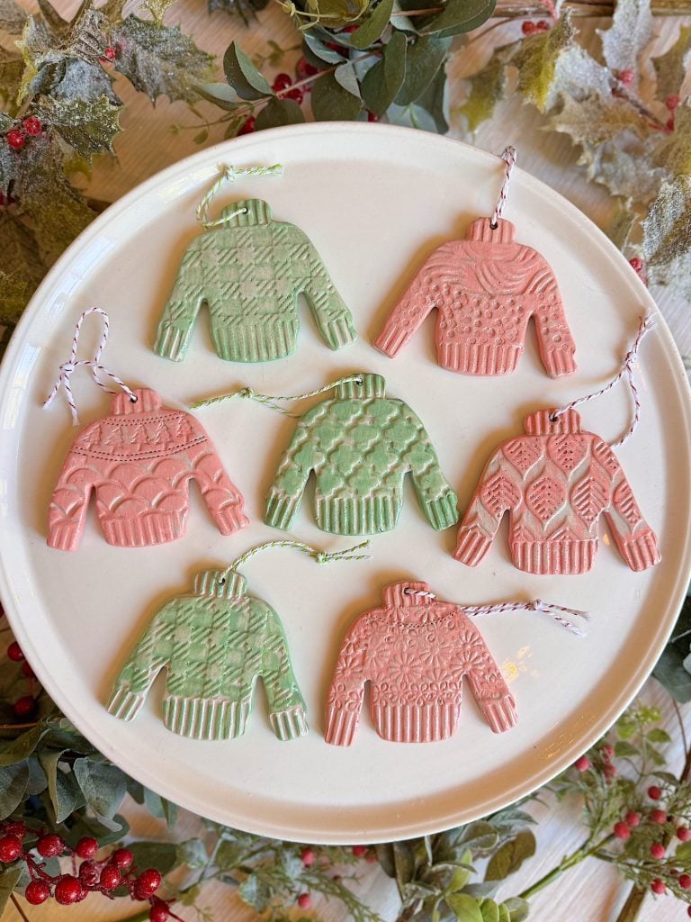 A white plate displays seven sweater-shaped ornaments in green and pink, each with a unique knit pattern and attached string, surrounded by festive greenery and berries.