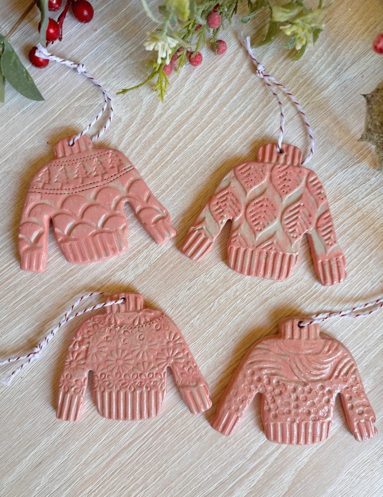 Four pink, sweater-shaped ornaments with textured patterns lie on a wooden surface, each hanging by a red-and-white string. Some greenery and berries are partially visible at the top.