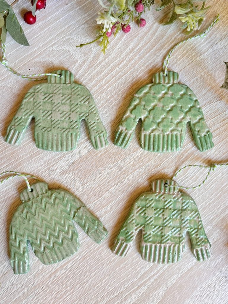 Four green ceramic ornaments shaped like sweaters with different textured patterns lie on a wooden surface; greenery decor is partially visible at the top.