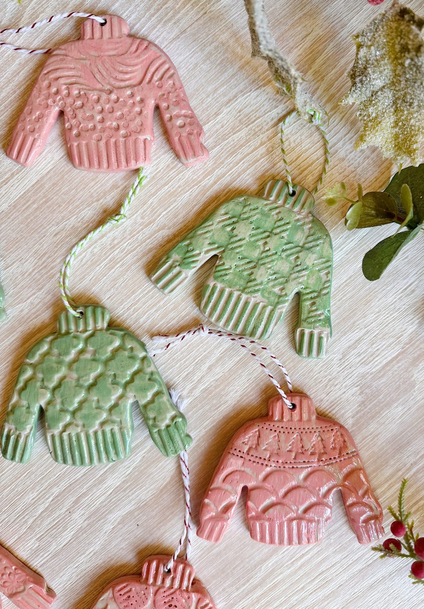 Assorted pink and green ceramic ornaments shaped like sweaters with textured patterns, arranged on a light wooden surface with twine loops attached.