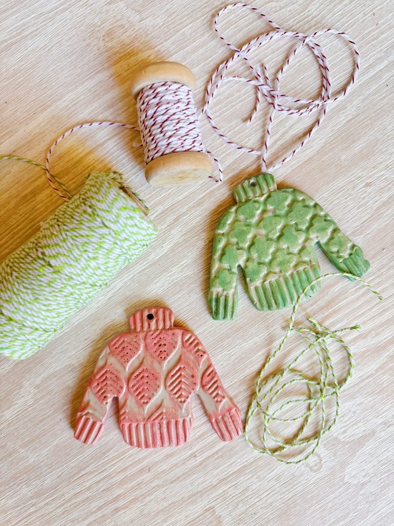 Two sweater-shaped ornaments, one red and one green, lie on a table next to spools of red-white and green-white twine. The ornaments have holes for hanging.