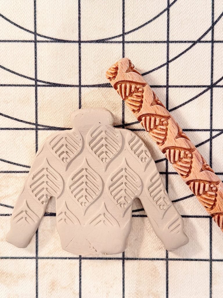 A clay sweater cutout with a leaf pattern sits beside a carved rolling pin on a gridded surface.