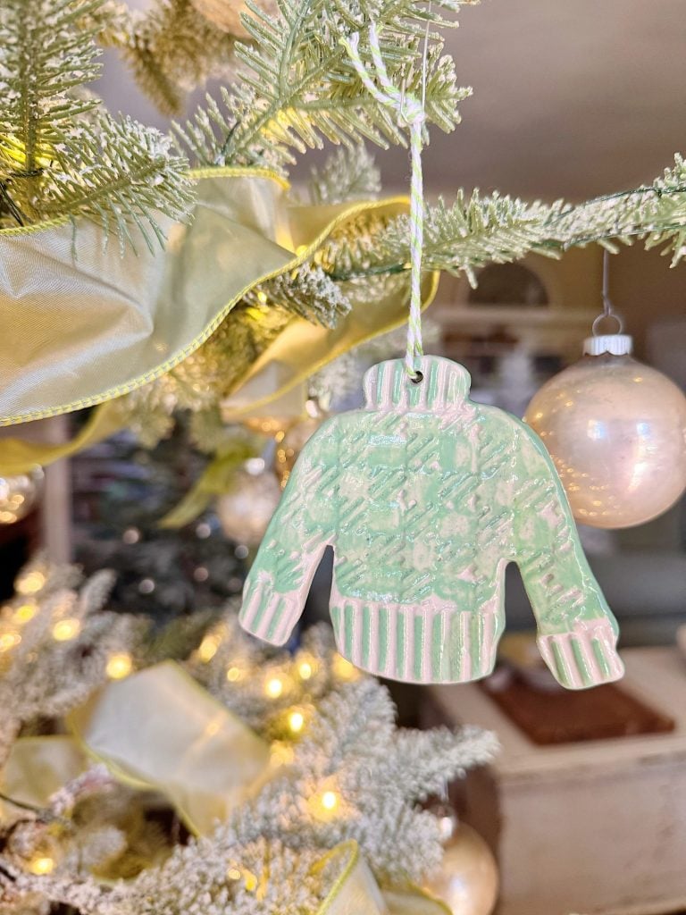 A green and white ceramic sweater ornament hangs on a Christmas tree decorated with lights, ribbon, and a round white ornament in the background.