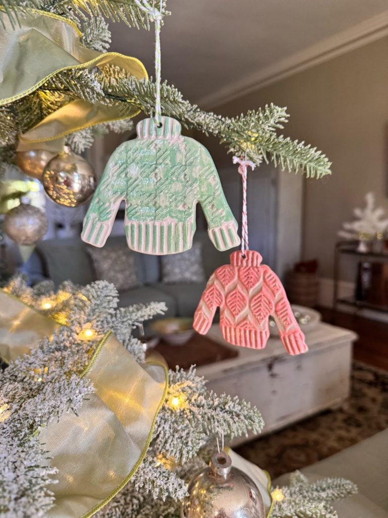 Two sweater-shaped ornaments, one green and one red, hang on a Christmas tree branch decorated with gold ribbon and lights, with a living room in the background.