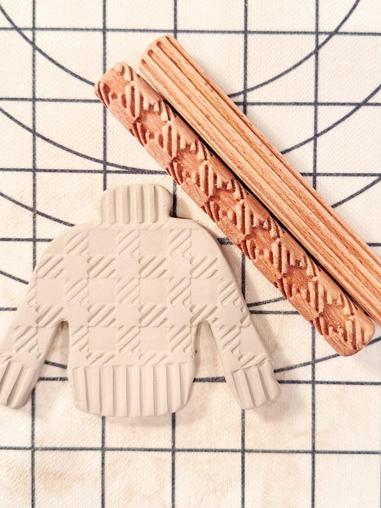 A clay cutout in the shape of a sweater with a textured pattern and two patterned wooden rollers on a grid-lined surface.