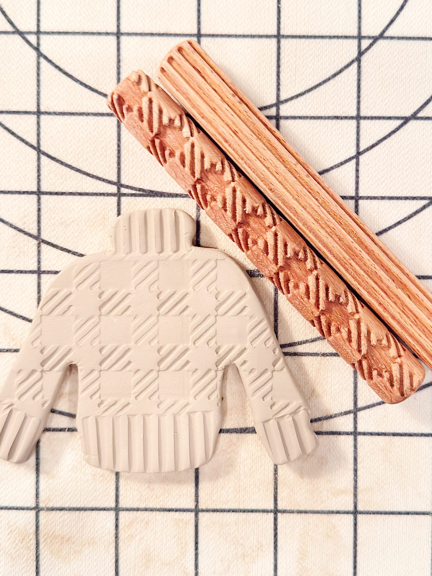 A clay cutout in the shape of a sweater with a textured pattern and two patterned wooden rollers on a grid-lined surface.