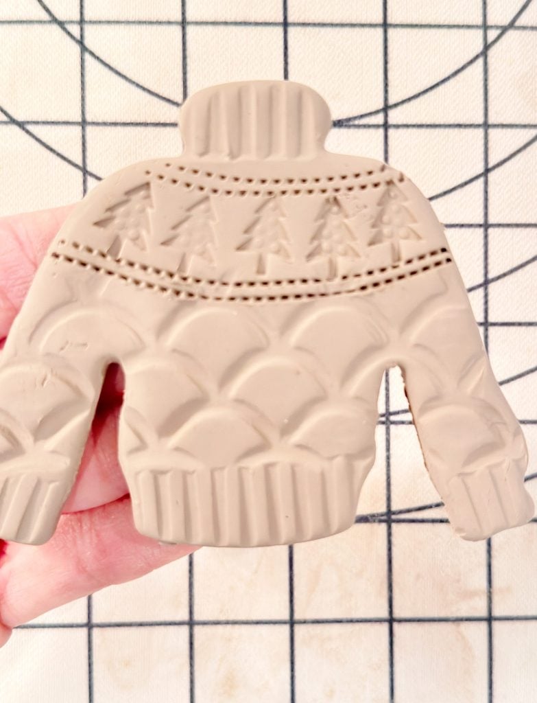 A hand holds a clay ornament shaped like a sweater with embossed patterns of trees, dots, and scallops against a gridded background.