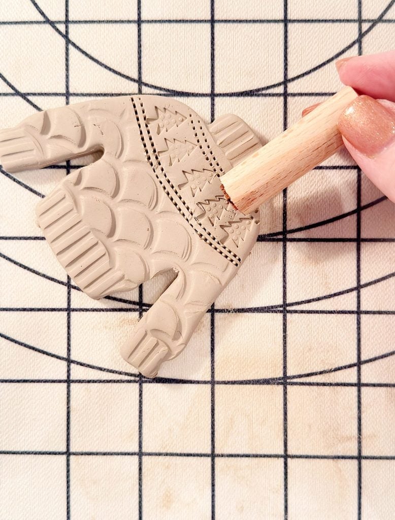 A hand uses a wooden tool to emboss a sweater-shaped clay ornament with a pattern on a gridded work surface.