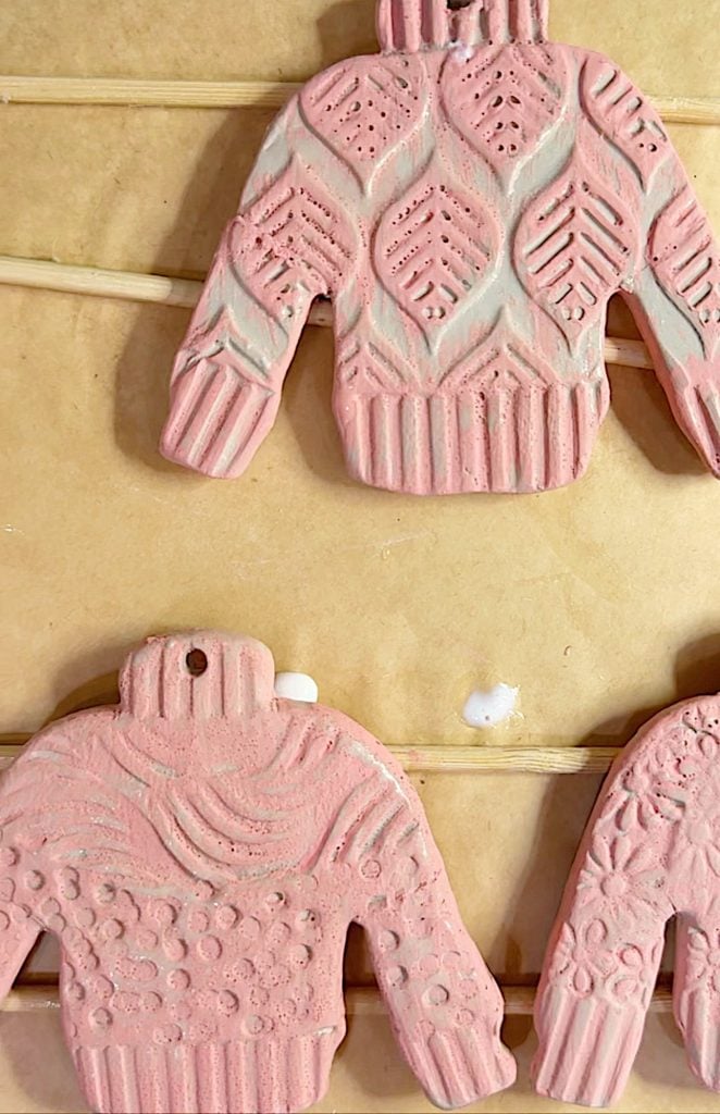 Three pink, ceramic sweater-shaped ornaments with textured patterns are displayed on a wooden rack.