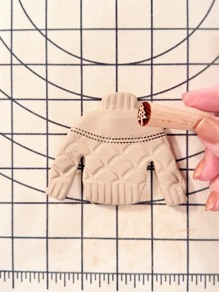 A hand presses a textured tool onto a clay cutout shaped like a sweater, which rests on a gridded surface.