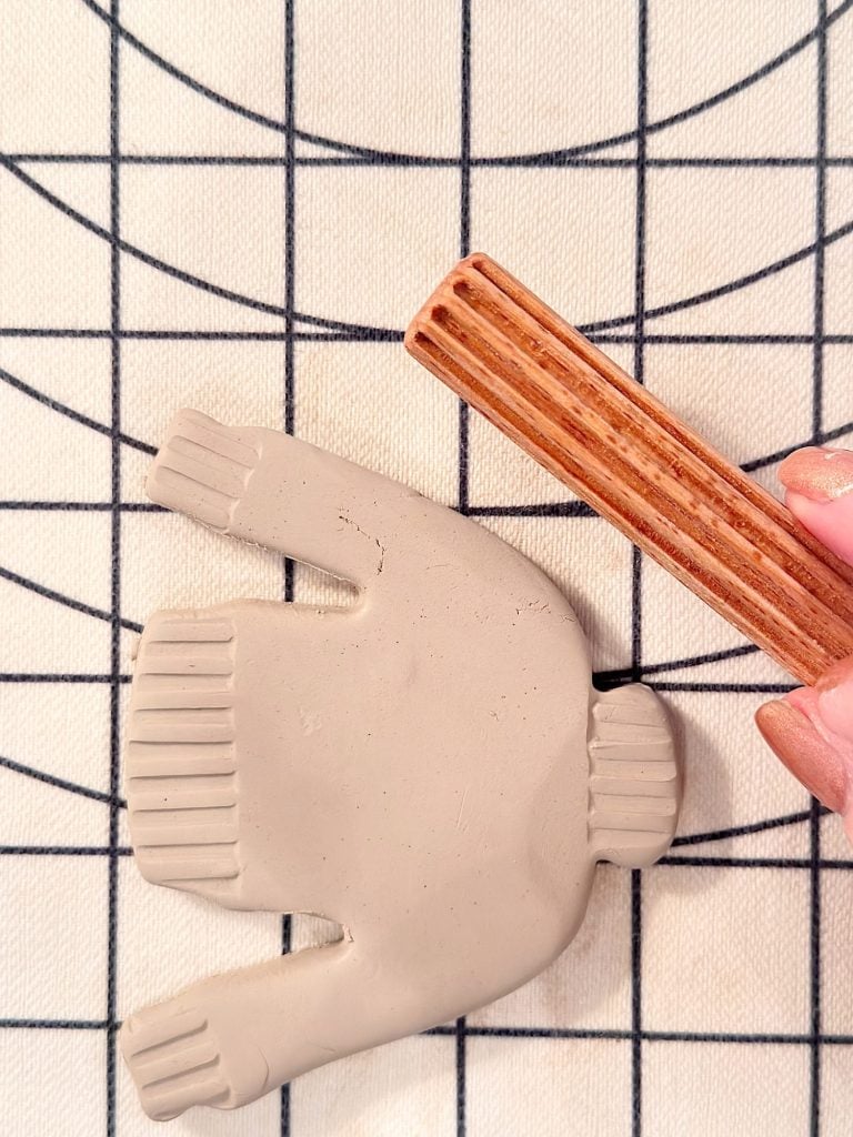 A hand uses a wooden tool to add textured lines to a clay cutout shaped like a sweater, placed on a gridded surface.