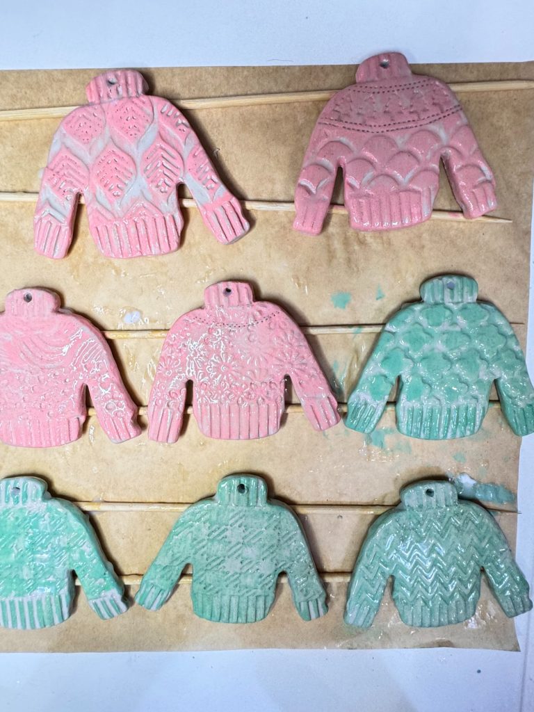 Eight decorated sweater-shaped cookies, four pink and four green, each with distinct knitted patterns, are arranged in two rows on parchment paper.