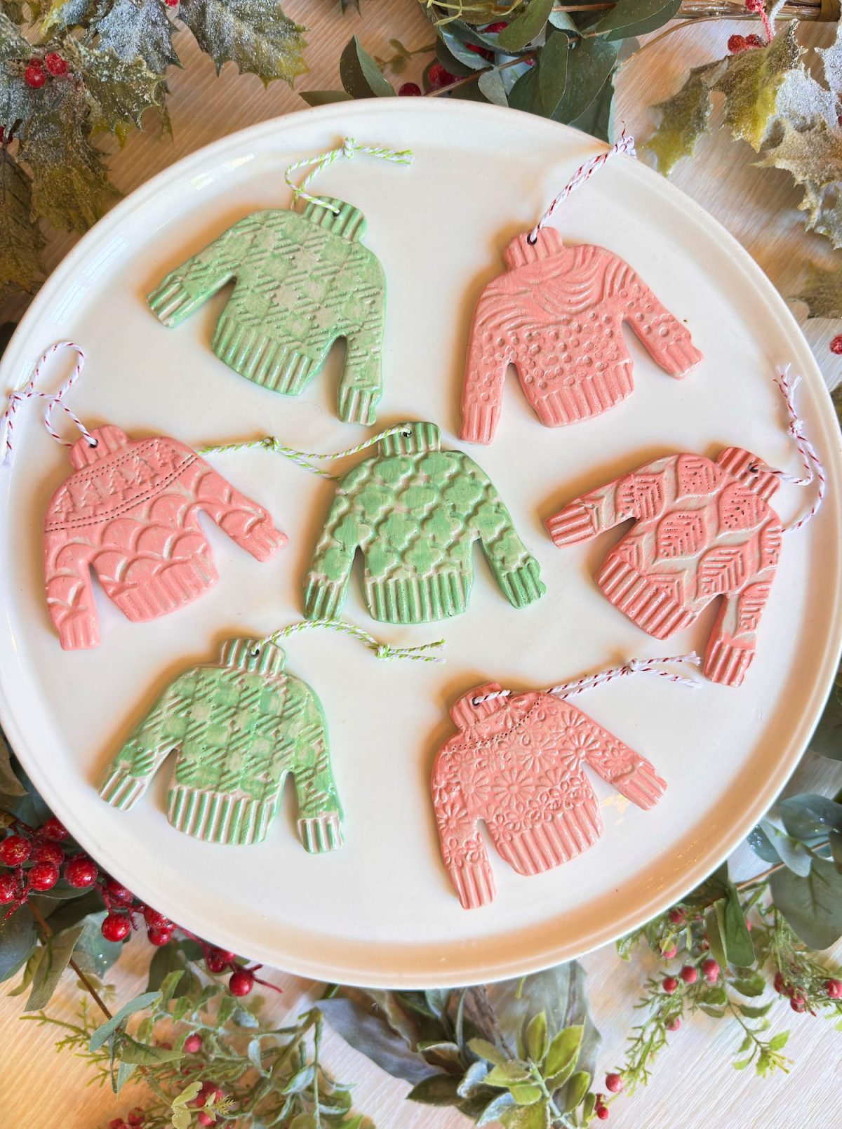 A white plate with six sweater-shaped ornaments in pink and green, featuring various textured patterns and attached with red and white or green and white string loops.