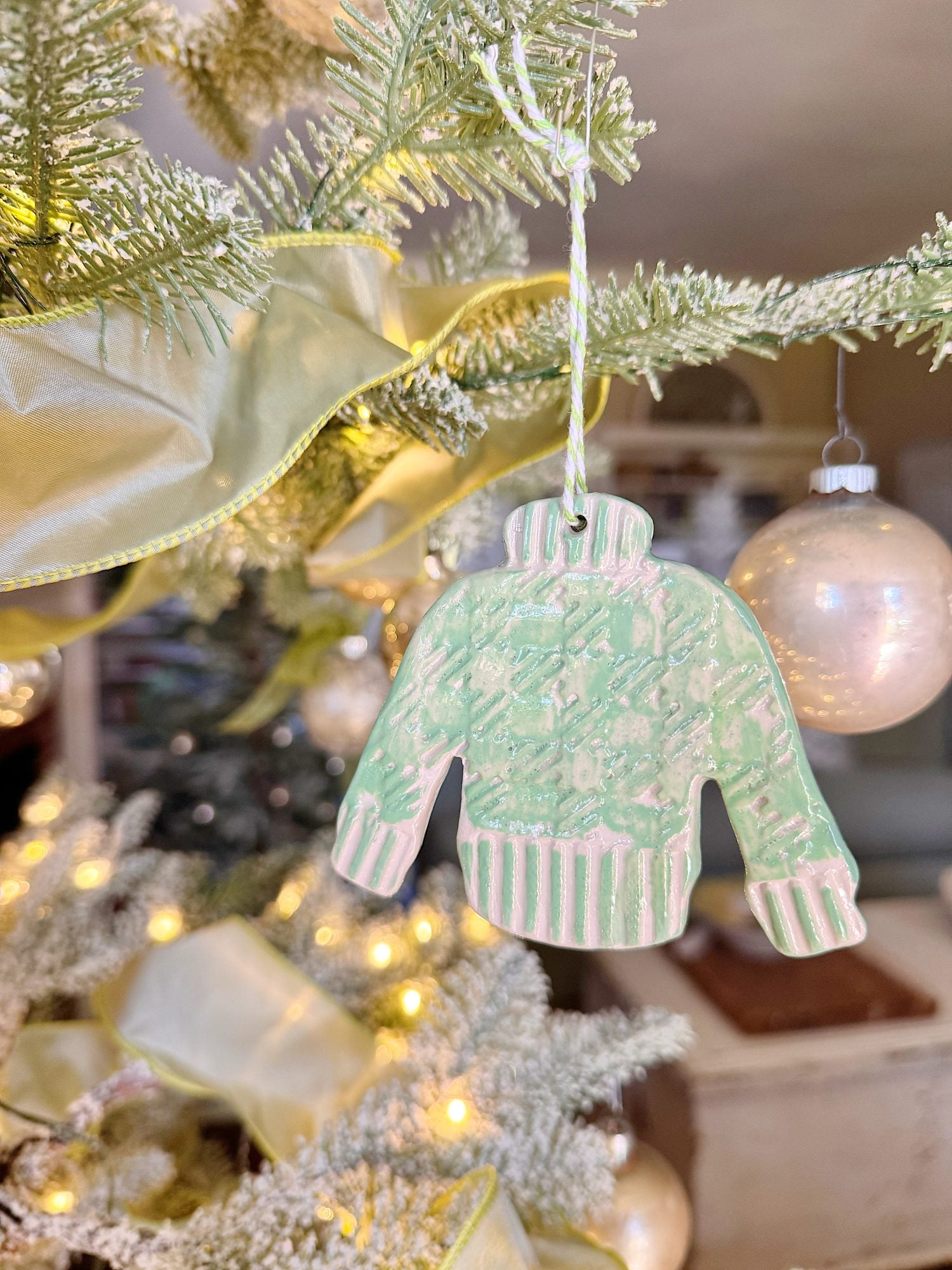 A green and white ceramic sweater ornament hangs on a Christmas tree decorated with lights, ribbon, and a round white ornament in the background.