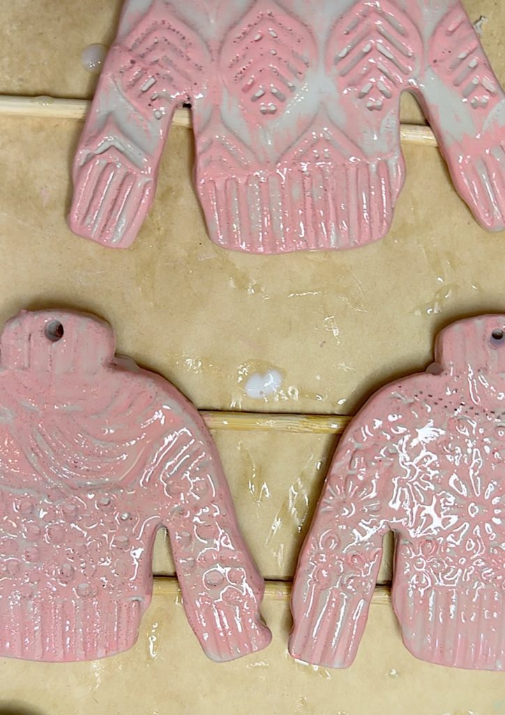 Three pink ceramic sweater ornaments with textured patterns hang on a rack, drying on a beige surface.