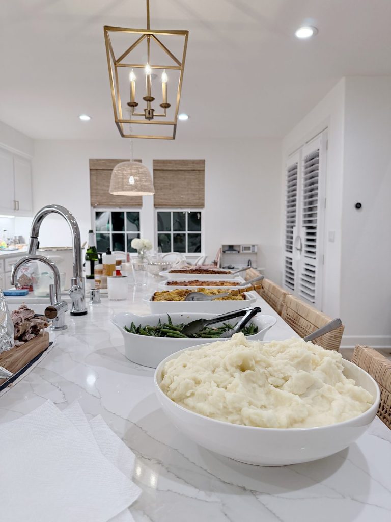 A kitchen countertop displays bowls of mashed potatoes, green beans, and other dishes, set under modern lighting in a bright, white kitchen accented with subtle Christmas decor.