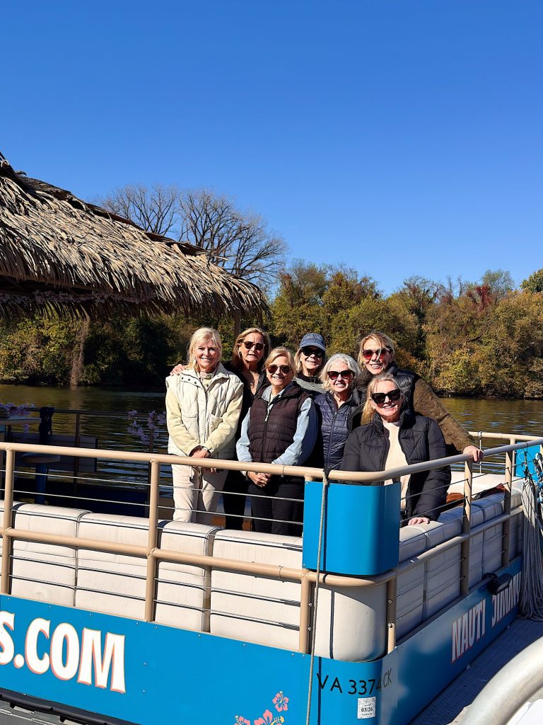 Seven women stand together and smile on a blue pontoon boat with a thatched roof, docked on a river, with trees and a clear sky in the background.