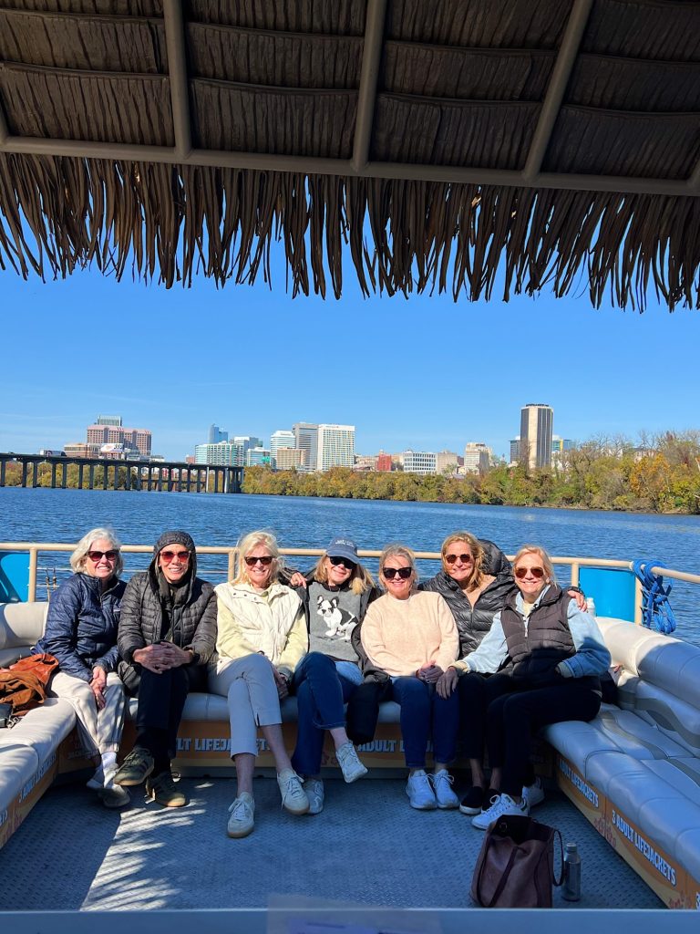 Seven women sit on the deck of a boat under a thatched roof, with a city skyline and blue sky visible across the river in the background.