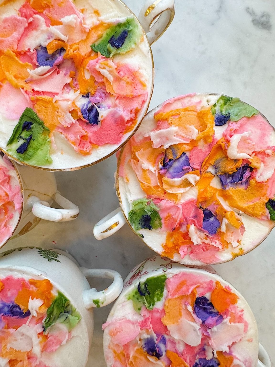 Several teacups filled with a colorful, textured mixture of pink, orange, green, and purple cream or frosting are arranged on a white marble surface&mdash;perfect inspiration for blog posts about creative dessert ideas.
