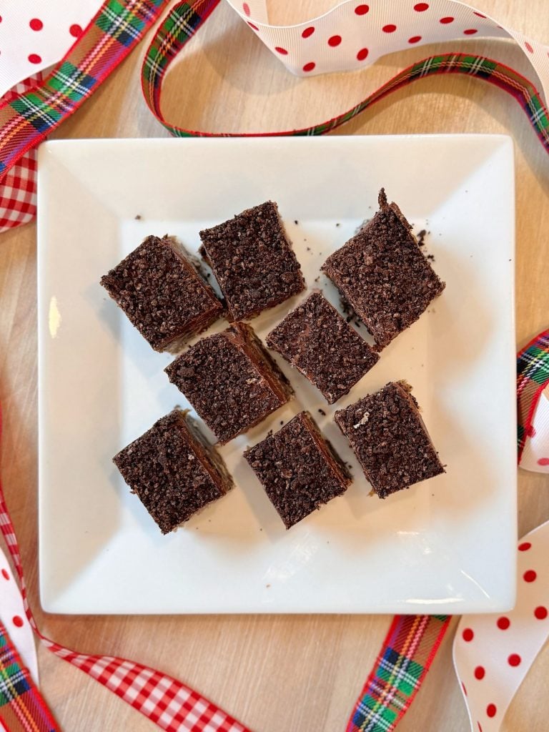 Nine small chocolate crumb squares are arranged in a 3x3 grid on a white square plate, with plaid and polka dot ribbons around the plate on a light wooden surface.