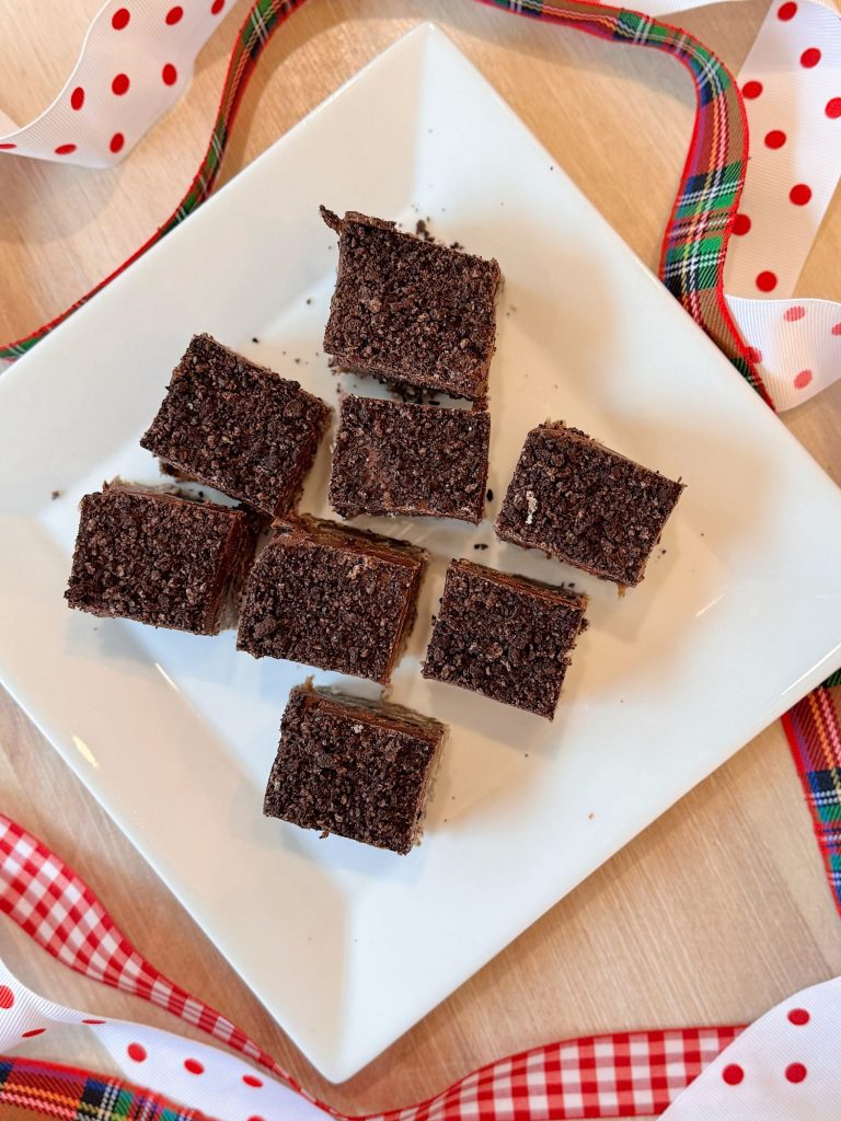 Nine chocolate crumb-topped dessert squares arranged on a white plate, surrounded by red, white, and plaid ribbon on a wooden surface.