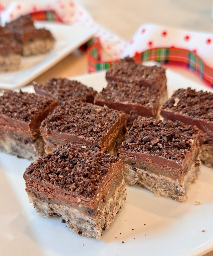 Squares of a layered dessert with a crumbly base, a chocolate topping, and a crunchy crumb layer on top, arranged on a white plate.