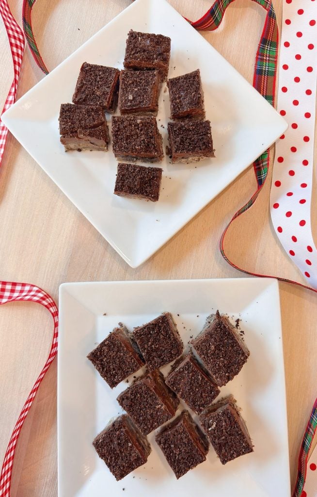 Two white square plates each hold nine chocolate dessert squares, with red and white decorative ribbons placed around them on a light wooden surface.