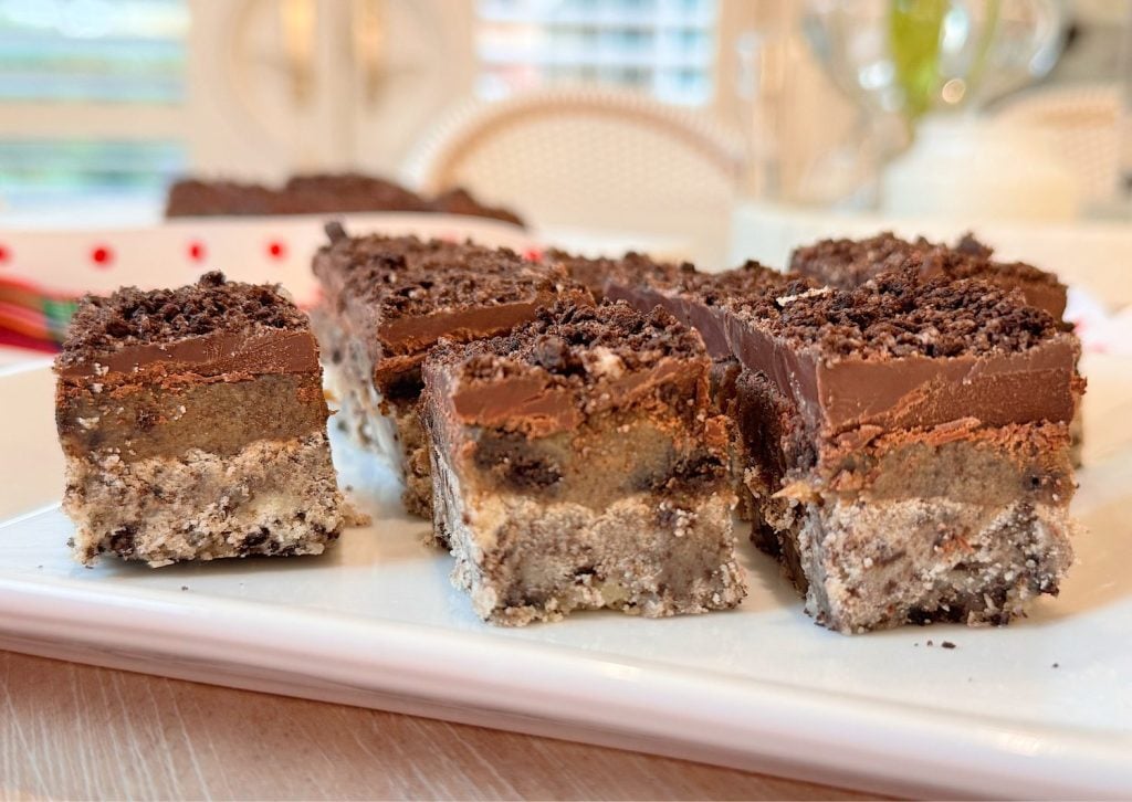 A close-up of dessert bars with three layers: a crumbly base, a thick middle layer, and a chocolate topping with crushed cookie pieces on top, arranged on a white plate.