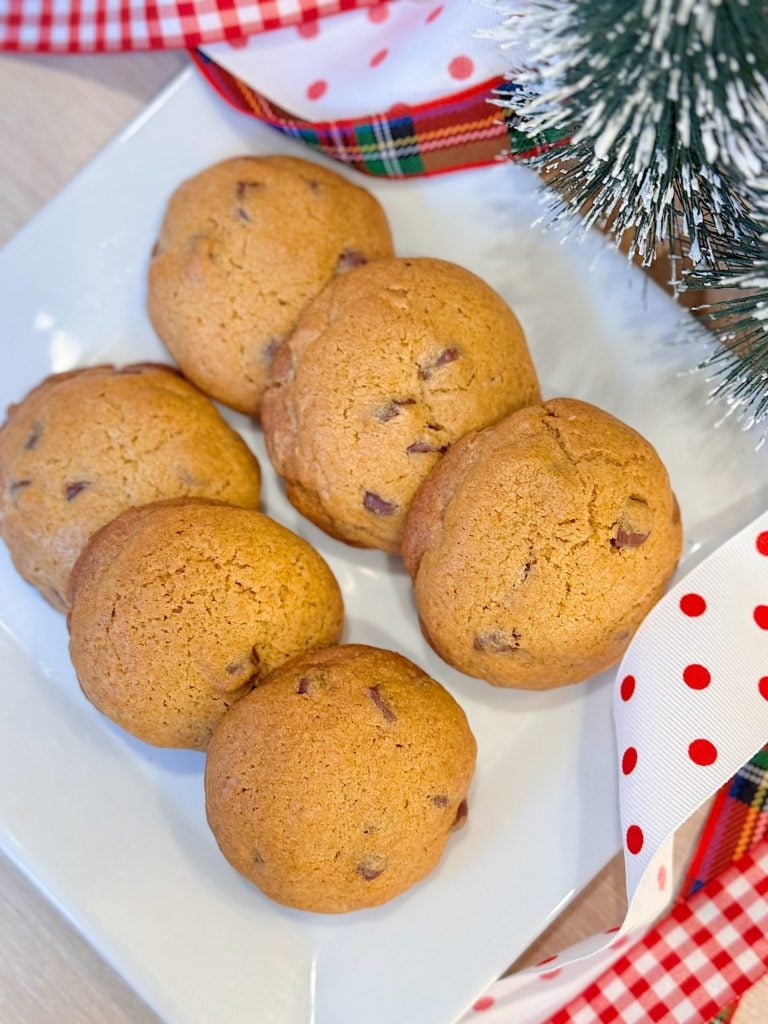 A white plate with seven chocolate chip cookies is placed on a table, decorated with red and polka dot ribbons and artificial pine branches.