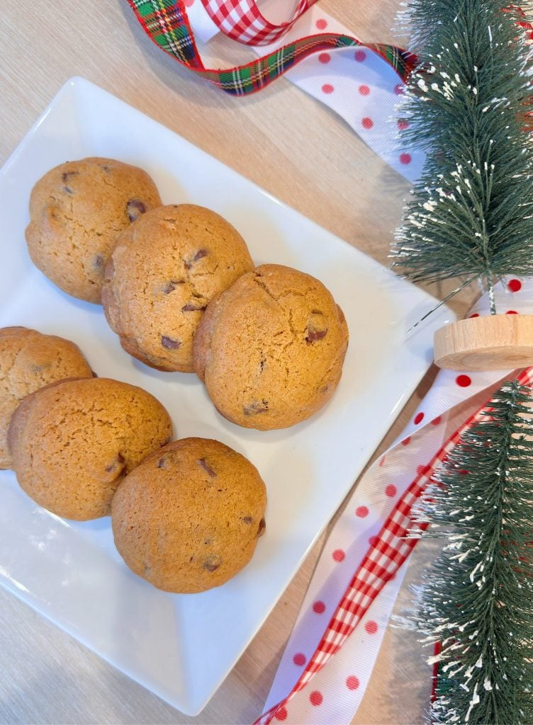 A white plate with six chocolate chip cookies next to a small artificial Christmas tree and red plaid and polka dot ribbons on a light wooden surface.