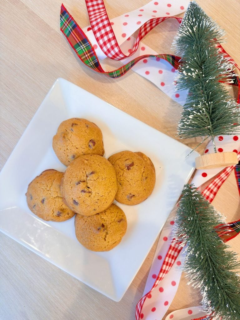 A white plate with five chocolate chip cookies is placed on a wooden surface, next to festive ribbons and artificial pine branches.