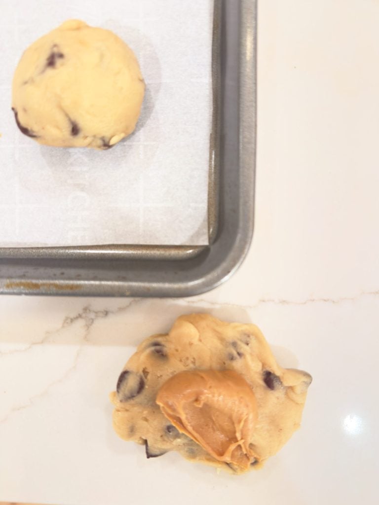 A baking tray with a ball of chocolate chip cookie dough and another flattened dough piece topped with a spoonful of peanut butter on a white counter.
