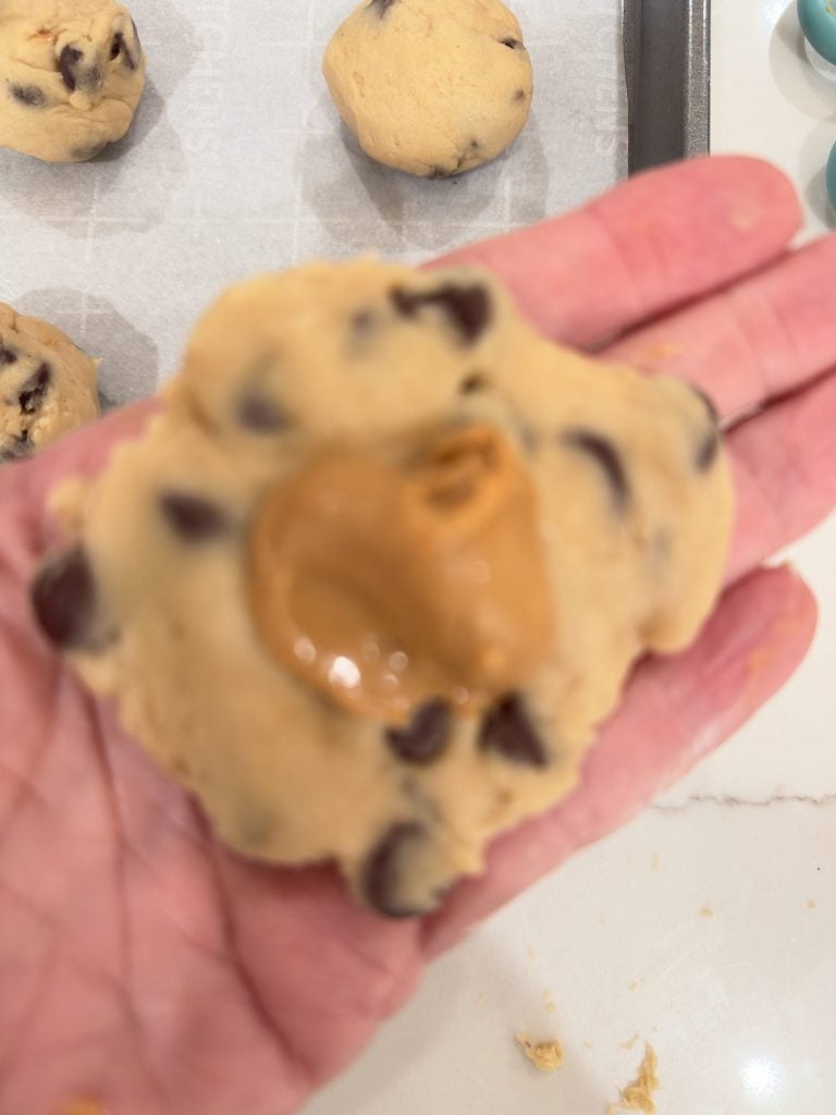 A hand holds a chocolate chip cookie dough ball with a dollop of peanut butter on top, ready to be shaped and baked.