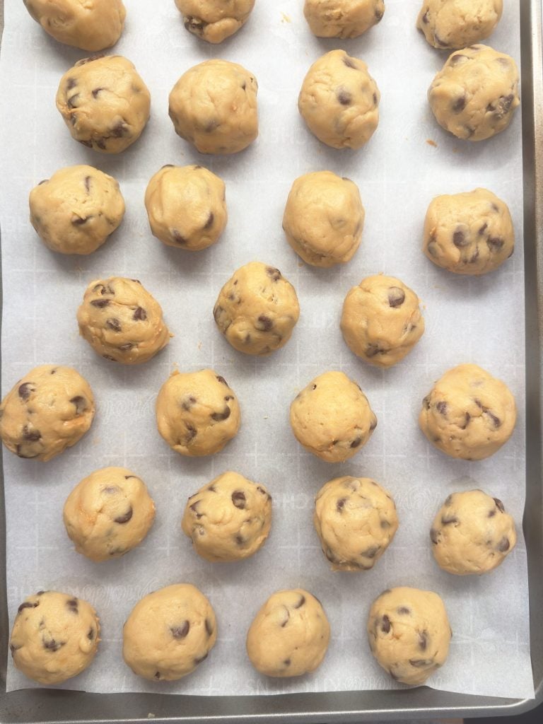 A baking tray lined with parchment paper holds evenly spaced balls of chocolate chip cookie dough, ready to be baked.