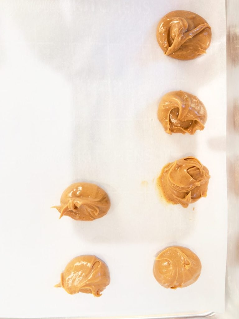 Six dollops of peanut butter are arranged on a parchment-lined baking sheet.