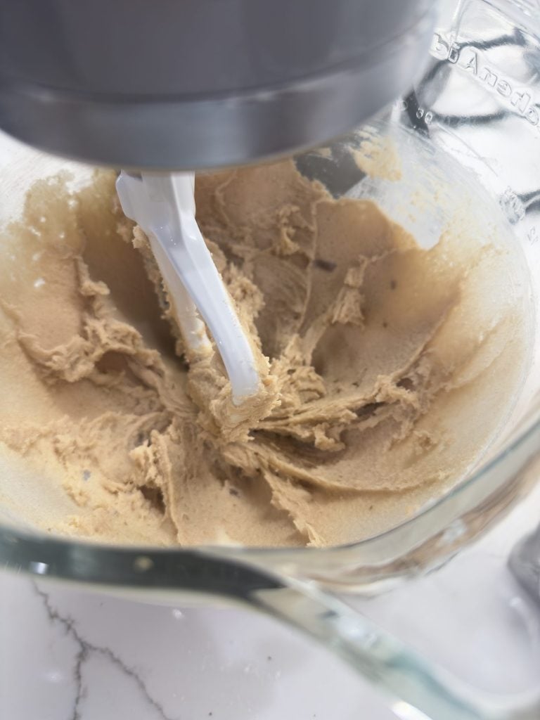 Stand mixer blending light brown cookie dough in a glass bowl on a white countertop.