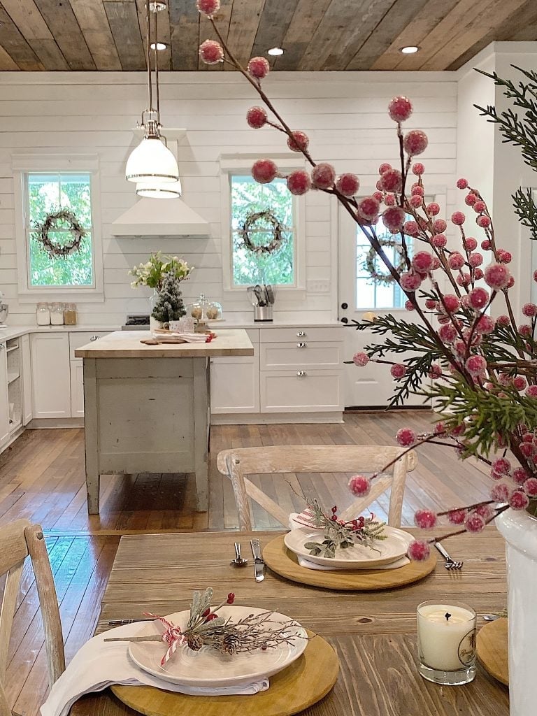 Bright farmhouse-style kitchen with white cabinets, rustic wood ceiling, and holiday-themed table setting with greenery and red berry accents.