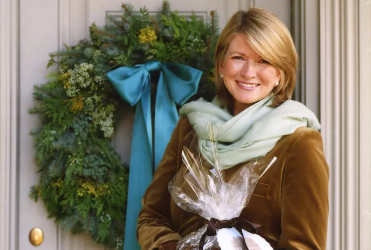 A woman wearing a brown jacket and light green scarf stands in front of a door with a holiday wreath, holding a wrapped gift.